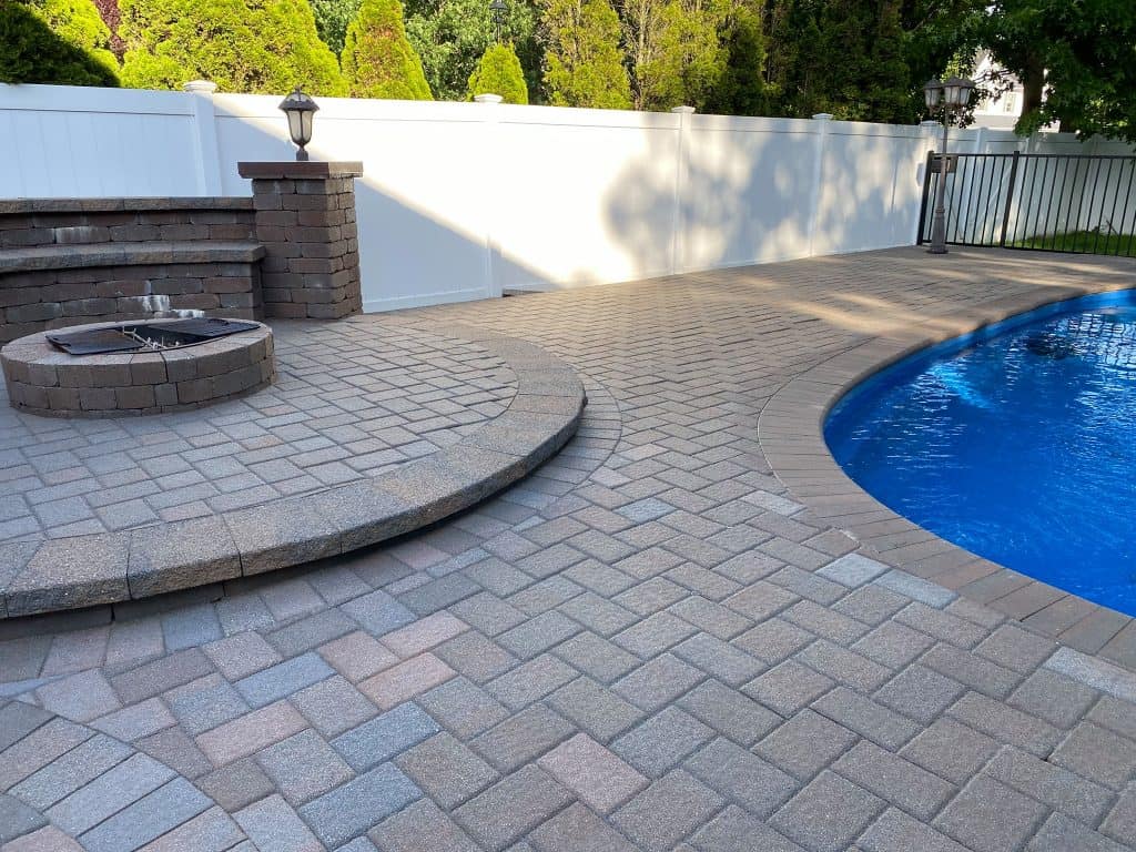 A backyard with a paved patio area next to a round blue swimming pool. There are brick steps leading up to a built-in fire pit surrounded by block seating. A white privacy fence lines the perimeter, and lush green trees are visible in the background, showcasing recent paver restoration in Nassau County.