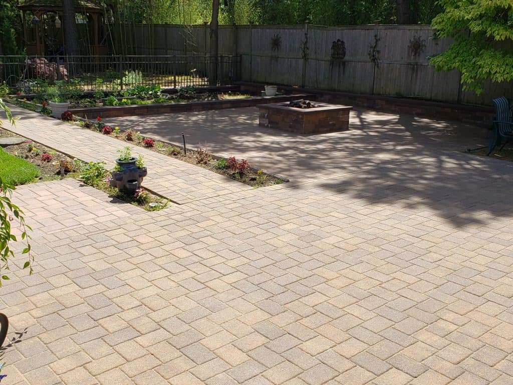 A backyard patio with light-colored pavers featuring a fire pit in the center and garden beds to the left. The space is bordered by a wooden fence, and surrounding trees provide partial shade. Paver restoration in Suffolk County has ensured that the path leading towards the fire pit area remains pristine.