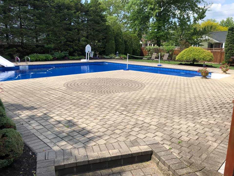 A spacious backyard features a large brick patio surrounding an in-ground swimming pool with a diving board, water slide, and a poolside basketball hoop. Lush greenery and tall trees add a natural touch to the area. For ongoing paver maintenance, consider expert Paver Sealing Nassau County services.