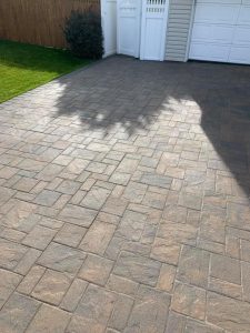 A driveway made of interlocking stone pavers in a light brown shade is in front of a white garage door. There is a shadow of a tree cast on the left side of the driveway, and a green lawn and a shrub are visible along a wooden fence in the background, showcasing excellent Paver Restoration Suffolk County.