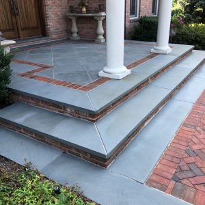 A multi-tiered porch with light gray stone tiles bordered by reddish-brown bricks features central columns supporting the roof. Brick steps with gray stone slabs lead to the entrance, and the pathway mirrors this design. For optimal maintenance, consider Paver Sealing Nassau County services.