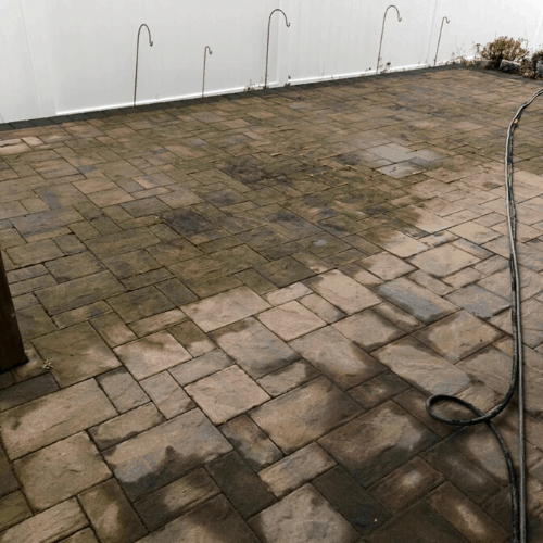 Image of a patio with stone pavers, some of which are darkened and wet from recent cleaning. A hose lies coiled on the ground with water splashing onto the pavers, indicating ongoing paver sealing in Nassau County. In the background, a white fence and several small garden hoses are visible.