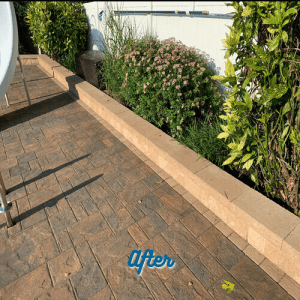 A landscaped area featuring a newly paved stone patio, bordered by a low retaining wall. Behind the wall are various green plants and flowering shrubs. Text at the bottom of the image reads "After." Paver sealing in Suffolk County ensures lasting beauty and durability for your outdoor spaces.