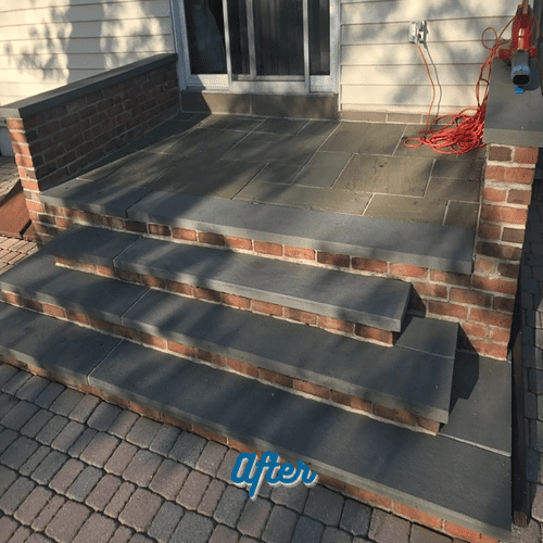 A newly constructed porch with four steps leading up to a door. The porch and steps are made of gray stone, with brick sides. A red extension cord and a blower are coiled up near the door, hinting at the recent Paver Restoration Nassau County work. The word "After" is written at the bottom of the image.