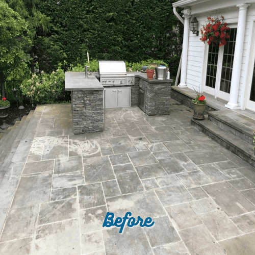 A patio with stone flooring and a built-in outdoor kitchen featuring a stone grill and countertop. The area is surrounded by greenery, and there are potted plants along with a flowering hanging basket near white double doors. "Before" is written on the ground—perfect for showcasing Paver Restoration Suffolk County services.