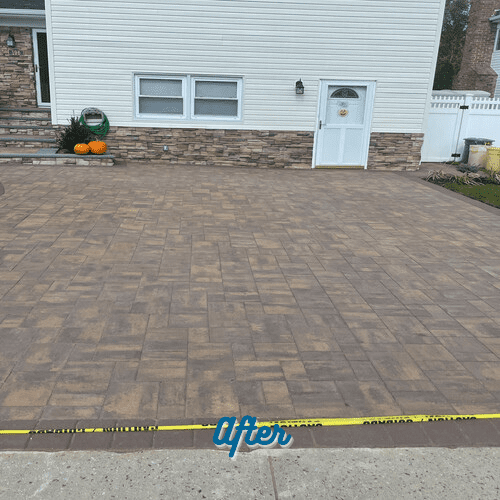 A newly completed driveway with a stamped concrete design graces the front of a white house with stone accents. A small flight of stairs leads to the front door, and there's a sideways door too. Yellow caution tape stretches across the driveway with a sign that says "After," suggesting recent paver repair in Nassau County.