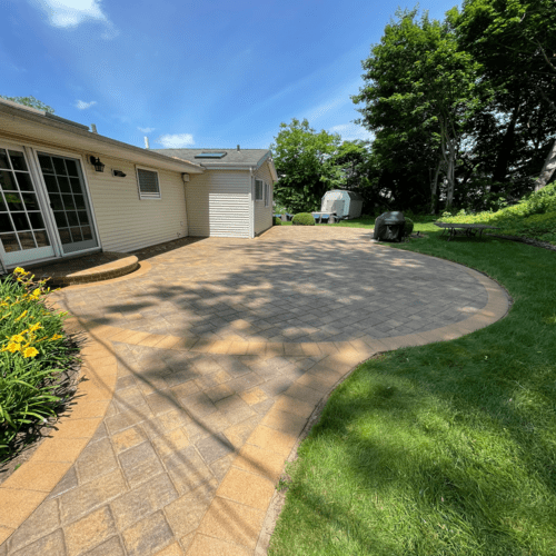 A backyard features a spacious patio paved with interlocking bricks in various shades. Adjacent to a white house with a sliding glass door, the patio is flanked by well-maintained grass, yellow flowers, a grill, and shaded by several trees under a blue sky. For longevity, consider paver sealing in Suffolk County.