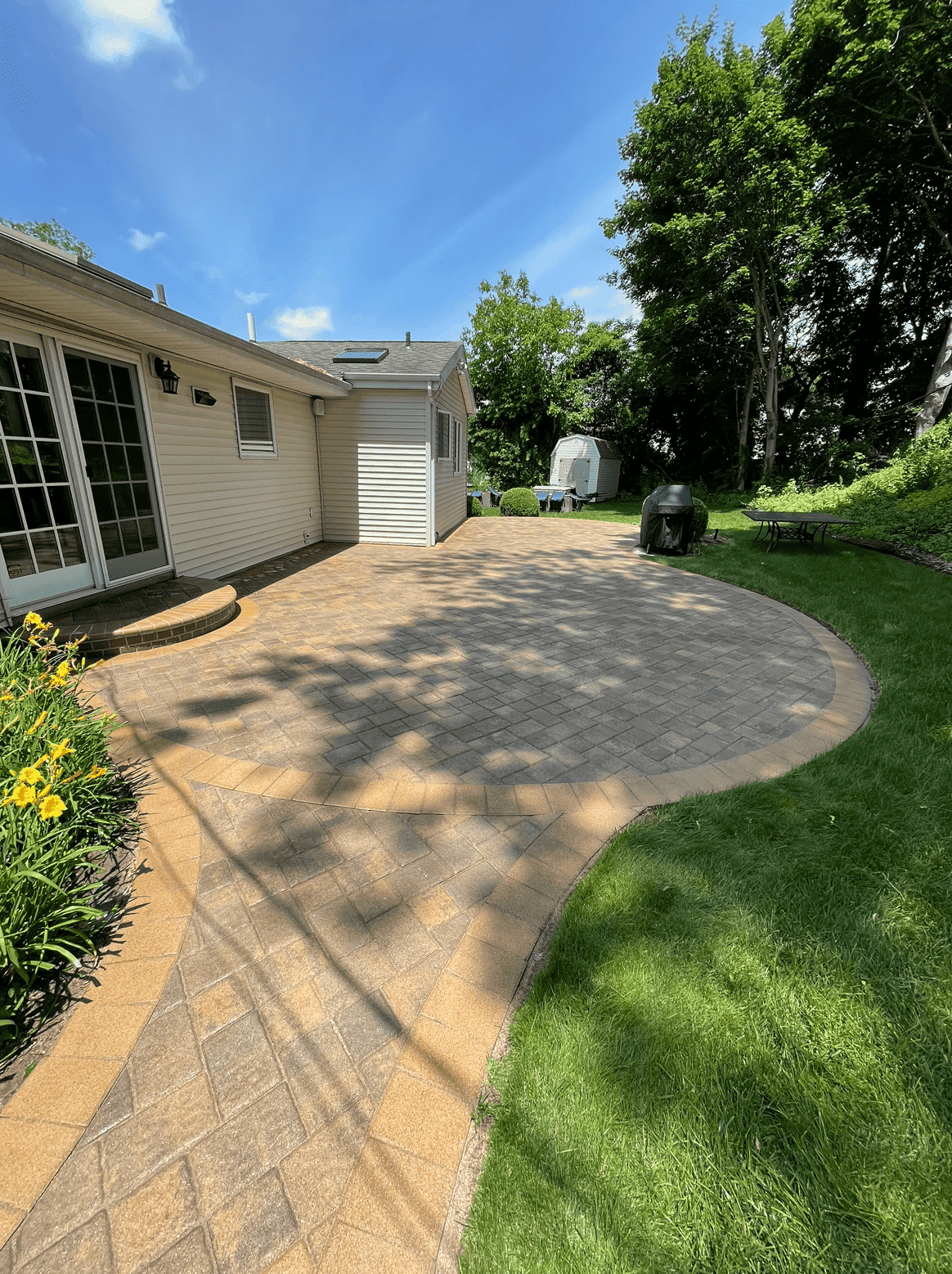 A backyard with a large stone patio adjacent to a house with beige siding and glass door. The herringbone-patterned patio, enhanced by expert paver sealing from Nassau County, is bordered by lush green grass and landscaped flower beds. Trees and shrubs surround the area, creating a peaceful outdoor space.