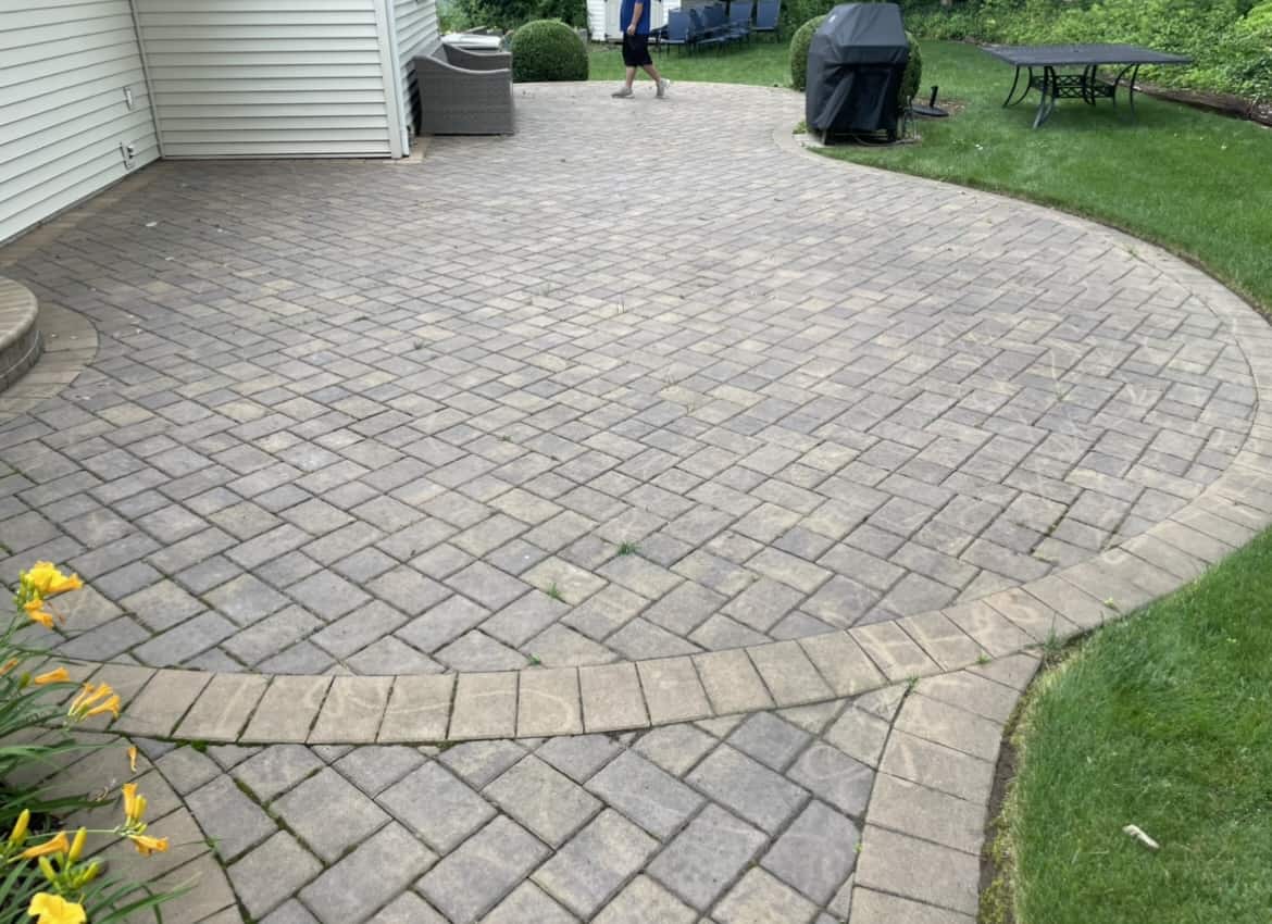 A spacious backyard patio with interlocking stone pavers, freshly maintained through Paver Sealing Nassau County, features a black grill to the right and a round table with chairs further back. There's a flowerbed with yellow flowers on the left and greenery surrounding the patio. A person is walking in the background.