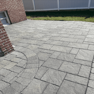 A newly constructed patio made of gray rectangular and square stones. The patio features a semi-circular design near a red brick wall and is bordered by a white fence. Green grass is visible at the edge of the patio, showcasing top-tier craftsmanship typical of Paver Restoration Suffolk County services.