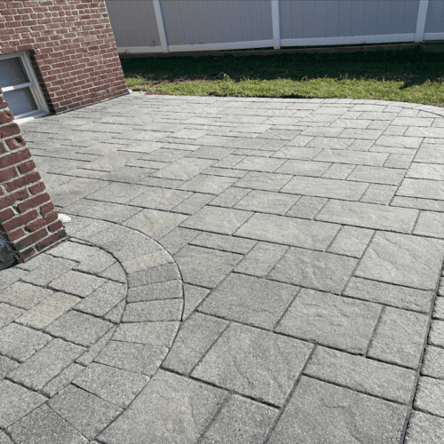 A newly constructed patio made of gray rectangular and square stones. The patio features a semi-circular design near a red brick wall and is bordered by a white fence. Green grass is visible at the edge of the patio, showcasing top-tier craftsmanship typical of Paver Restoration Suffolk County services.