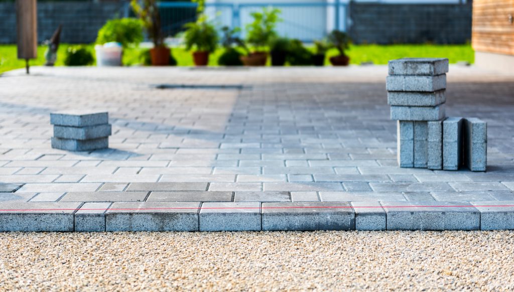 A partially completed patio with neatly arranged gray paving stones. A stack of stones is on the right, and a smaller stack is on the left. There is a gravel base in the foreground with a red string line for alignment. Potted plants are visible in the background, awaiting paver restoration Nassau County services.