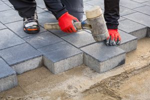 A person with red gloves is placing gray concrete pavers into a sand base, using a rubber mallet to adjust their position. They are wearing black pants and sneakers. The scene, seemingly involving paver repair in Nassau County, appears to be part of a construction or landscaping project.