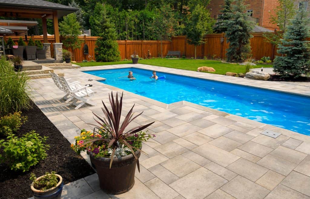 A backyard with a tiled patio area, a swimming pool with two children swimming, and surrounding greenery. There are lounge chairs near the pool, a table with chairs under a gazebo, and potted plants enhancing the landscape. Paver sealing in Nassau County ensures the wooden fence enclosing the area stays pristine.