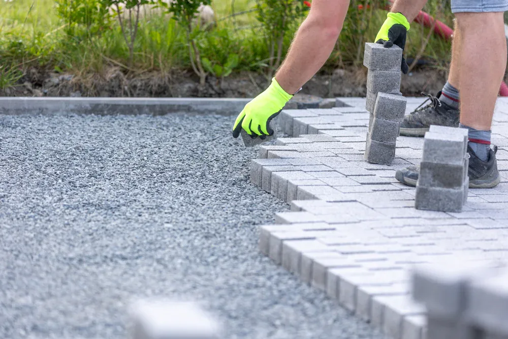 A person wearing neon yellow gloves and shorts is laying gray pavers Long Island-style on a gravel base while constructing a patio or walkway outdoors in NY.