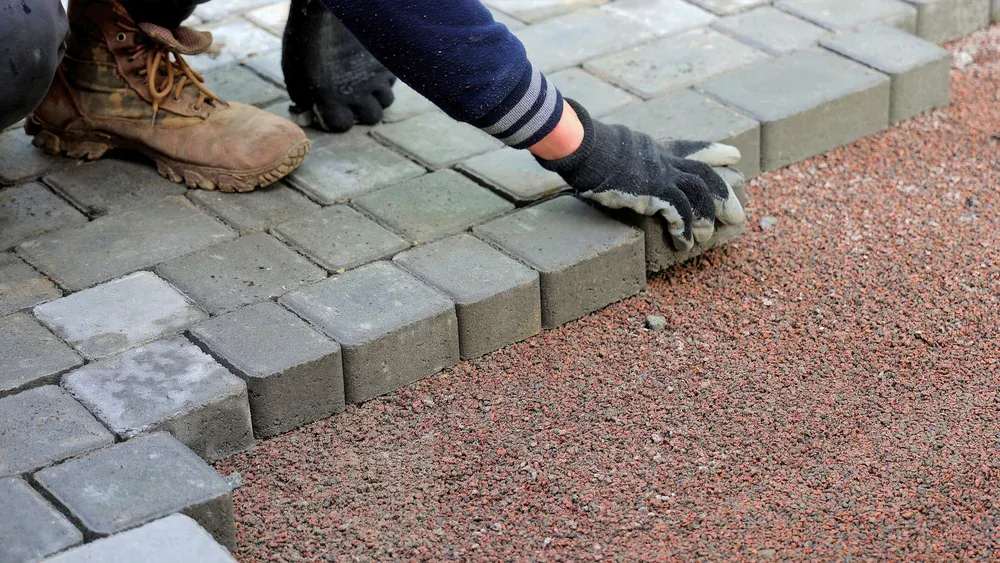 A person wearing work gloves and boots is laying rectangular gray pavers Long Island style on prepared ground, arranging them in neat rows over a layer of reddish gravel in NY.