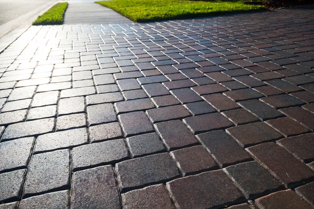 Close-up of a clean, sunlit sidewalk crafted from neatly arranged rectangular and square brick pavers Long Island, NY, with green grass and a concrete curb visible in the background.