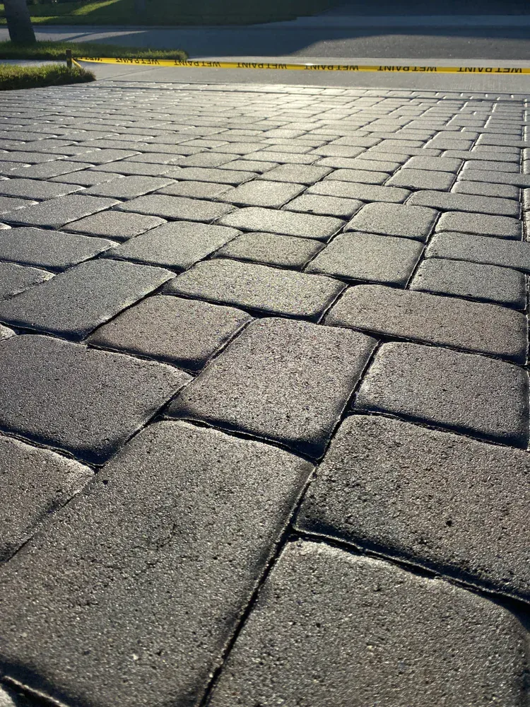 Close-up view of sunlit stone pavers forming a walkway or driveway, with a yellow caution tape visible in the background, indicating a restricted or under-construction area.
