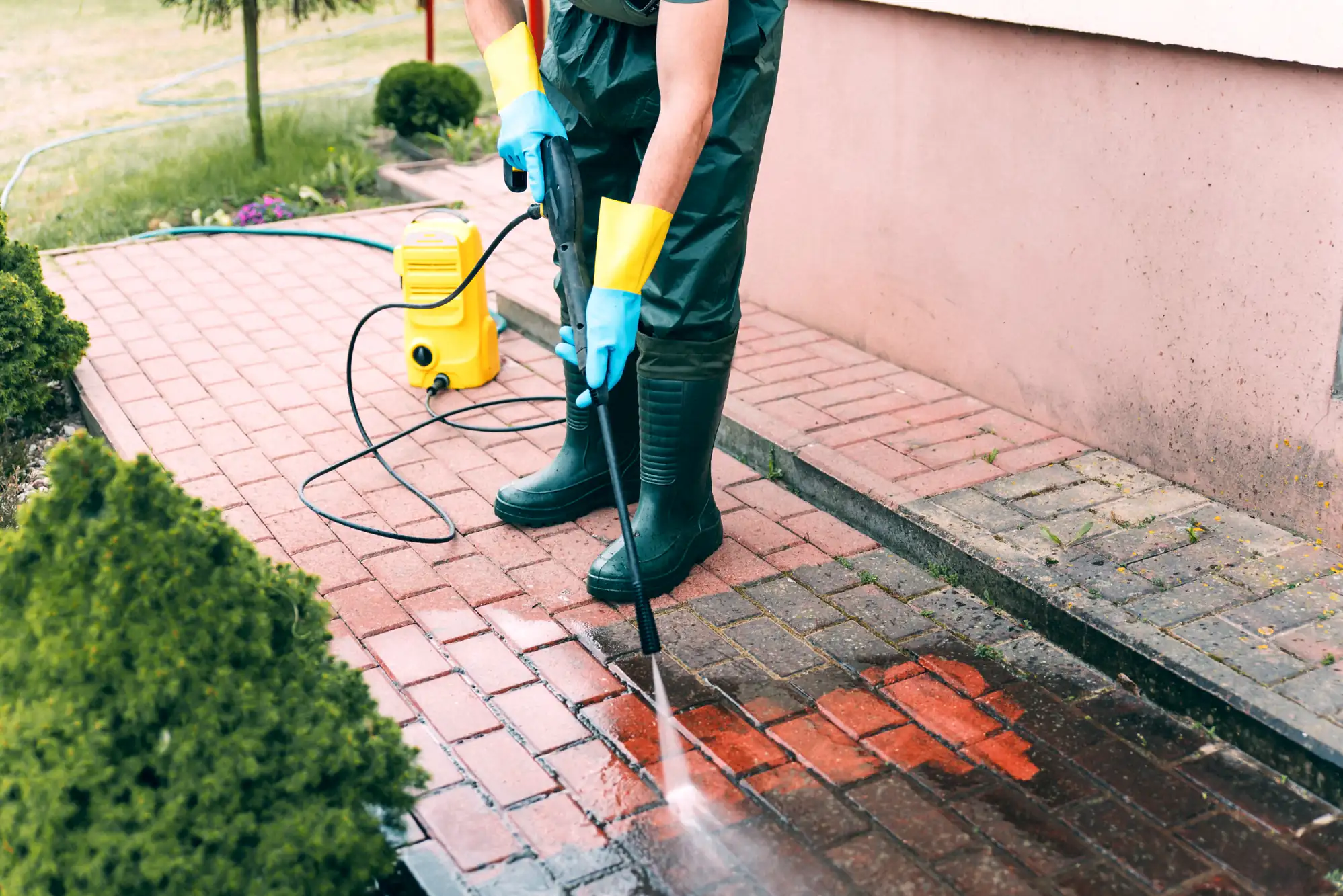 Person in boots and gloves power washing a red brick sidewalk with a yellow pressure washer outside.