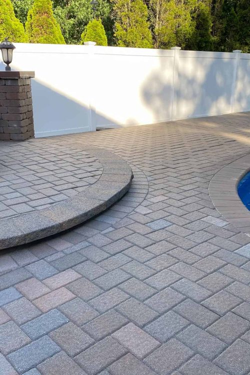 A backyard with a paved patio area next to a round blue swimming pool. There are brick steps leading up to a built-in fire pit surrounded by block seating. A white privacy fence lines the perimeter, and lush green trees are visible in the background, showcasing recent paver restoration in Nassau County.