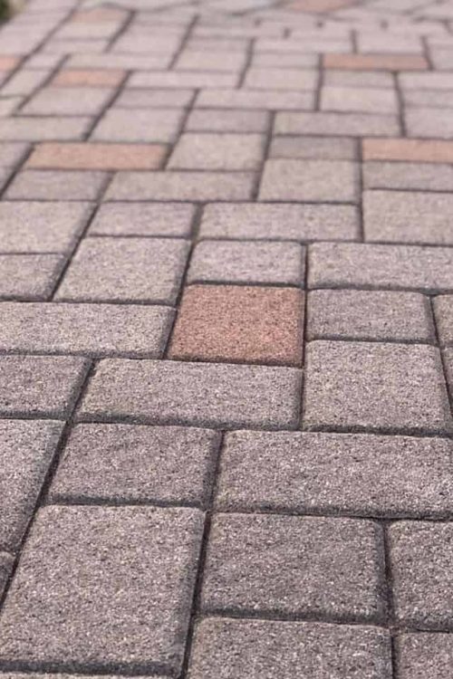A close-up of a neatly arranged, multicolored brick pathway shows the results of expert paver restoration in Nassau County. The bricks are primarily shades of gray and light pink, forming a herringbone pattern. The path extends into the distance, slightly curving, with patches of grass visible on the side.