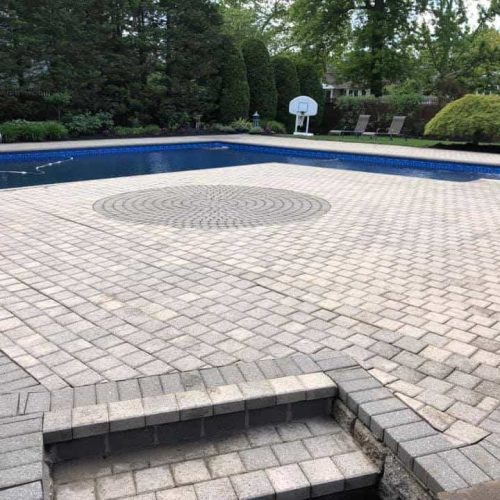 A backyard pool surrounded by a paved patio with a decorative circular pattern at the center. There is a pool slide and a basketball hoop at one end of the pool. Steps lead down from a wooden deck onto the patio, which benefits from professional Paver Sealing Nassau County services. Trees and shrubs are visible in the background.