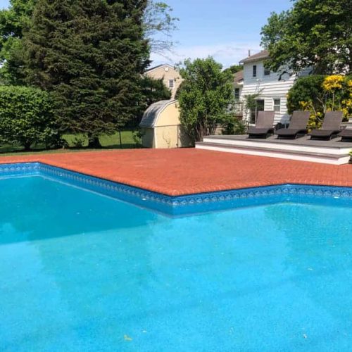 A backyard swimming pool with clear blue water, surrounded by a red brick deck. In the background, there's a small deck with four brown lounge chairs and a green hedge. Trees and houses are visible beyond the hedge, showcasing excellent paver restoration in Nassau County under a clear and sunny sky.