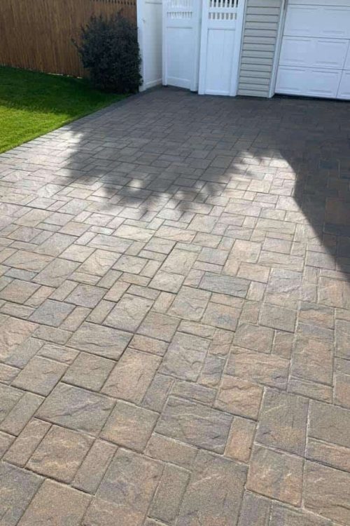 A driveway made of interlocking stone pavers in a light brown shade is in front of a white garage door. There is a shadow of a tree cast on the left side of the driveway, and a green lawn and a shrub are visible along a wooden fence in the background, showcasing excellent Paver Restoration Suffolk County.