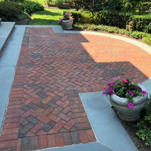 A patio featuring a neat herringbone pattern made from red and brown bricks, bordered by large grey tiles. There are two large white planters with bright pink and purple flowers at each end of the patio. Lush green grass and shrubs surround the area, showcasing expert paver restoration in Suffolk County.