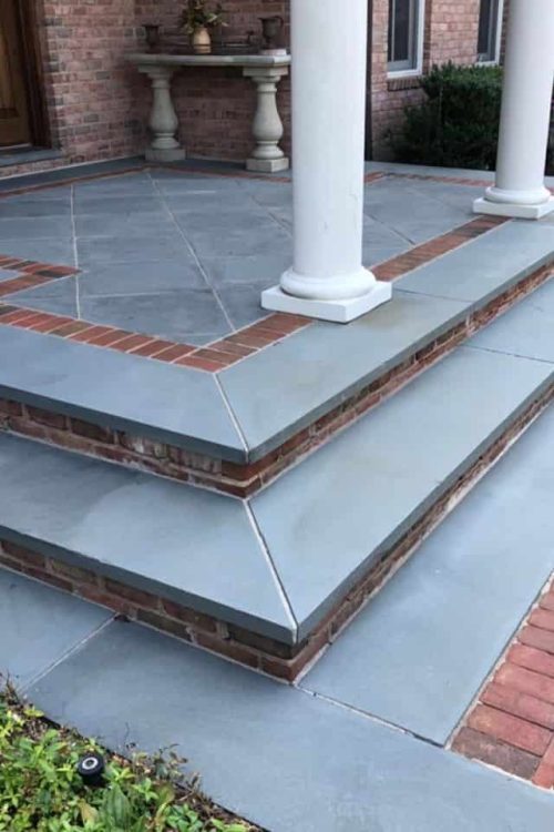 A multi-tiered porch with light gray stone tiles bordered by reddish-brown bricks features central columns supporting the roof. Brick steps with gray stone slabs lead to the entrance, and the pathway mirrors this design. For optimal maintenance, consider Paver Sealing Nassau County services.
