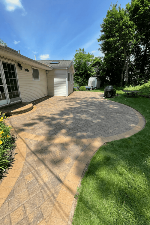 A backyard with a large stone patio adjacent to a house with beige siding and glass door. The herringbone-patterned patio, enhanced by expert paver sealing from Nassau County, is bordered by lush green grass and landscaped flower beds. Trees and shrubs surround the area, creating a peaceful outdoor space.