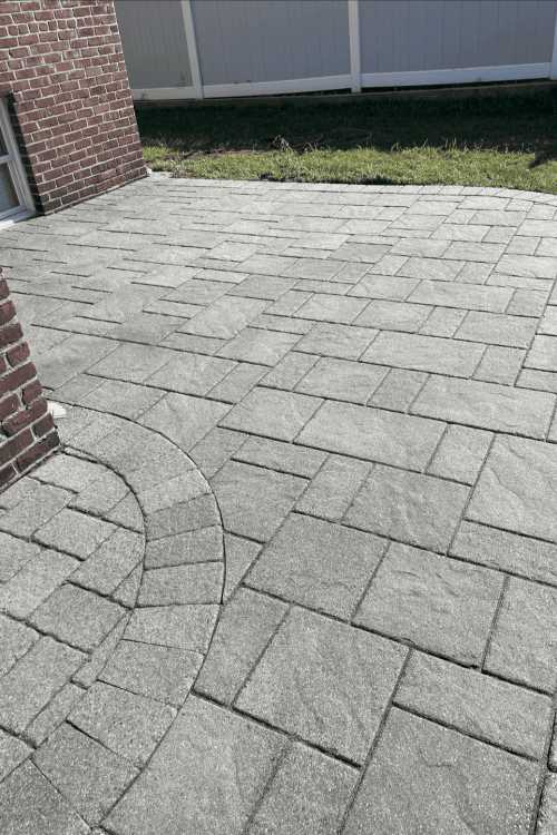 A paved patio with gray stone tiles in a geometric pattern is adjacent to a red brick wall. A small grassy area and a white fence are visible in the background, showcasing exquisite work ideal for Paver Sealing Nassau County.