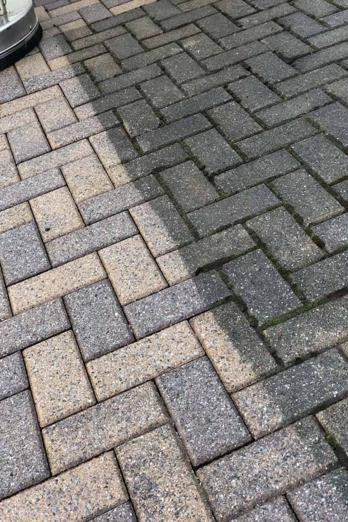 A brick driveway is shown, with a clear contrast between the left side, which is clean and light-colored, and the right side, which is dark and dirty. A cleaning machine is partially visible in the upper left corner, halfway through its task of paver restoration in Nassau County.
